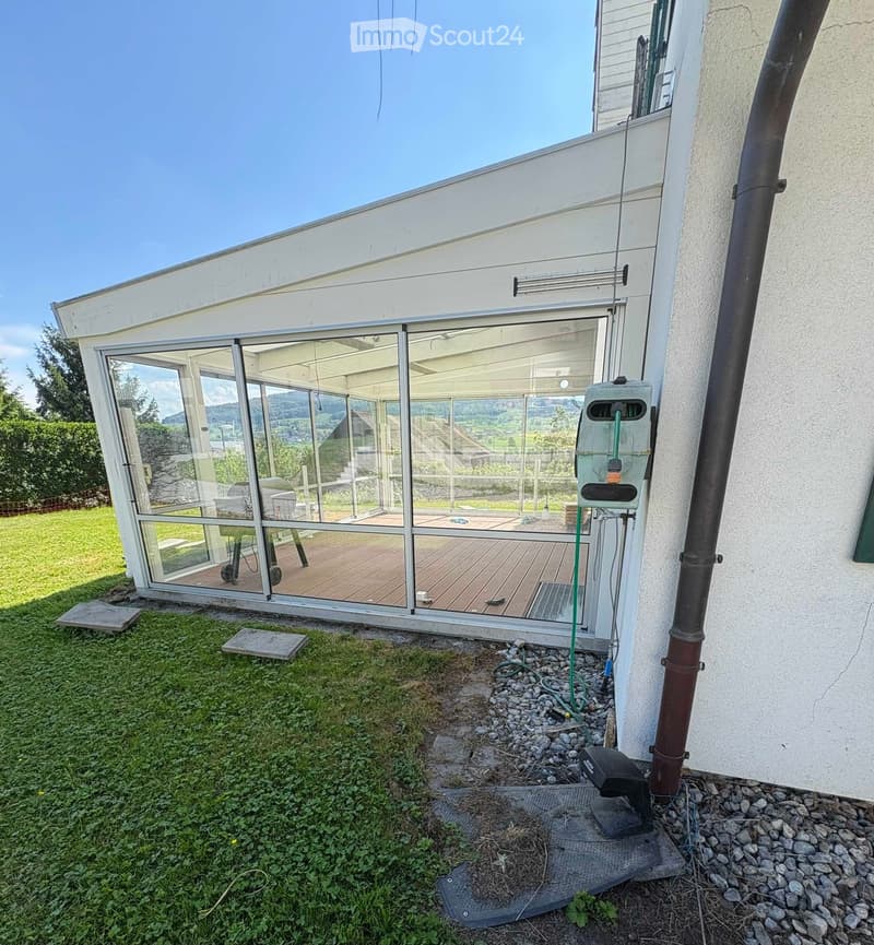 A modern glass-enclosed patio attached to a house, with wooden flooring, a view of the surrounding landscape, and some outdoor equipment visible near the entrance.