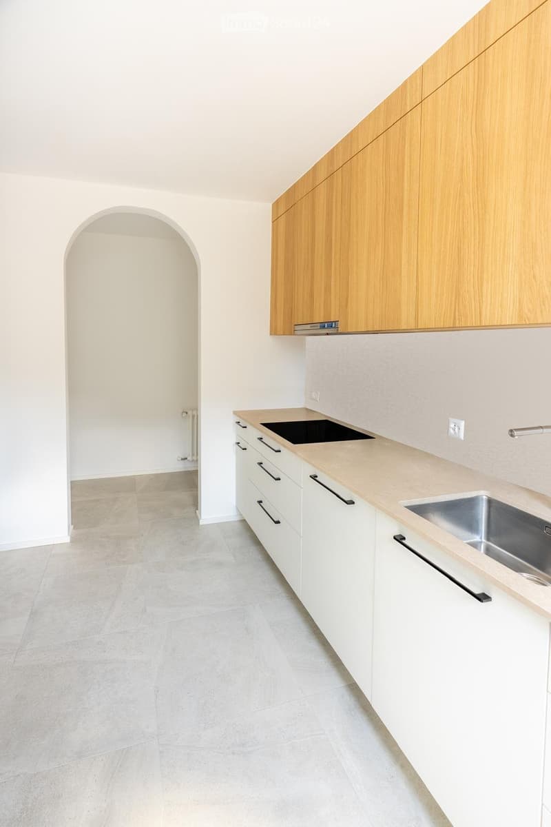 Modern kitchen with light wooden cabinets, white countertops, a sink, and a tiled floor. The space is clean and well-lit with an arched doorway leading to another room.