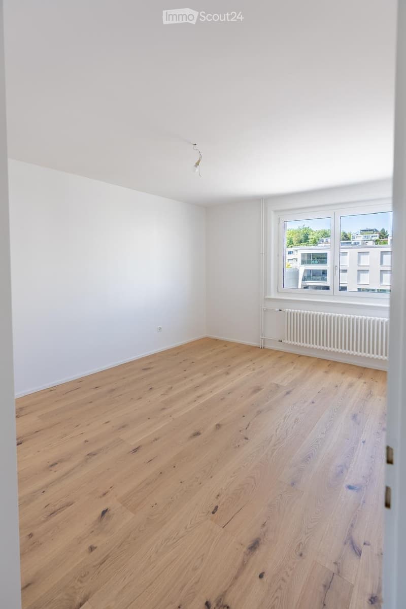 Empty room with light wooden flooring, white walls, and a large window with a radiator below it.