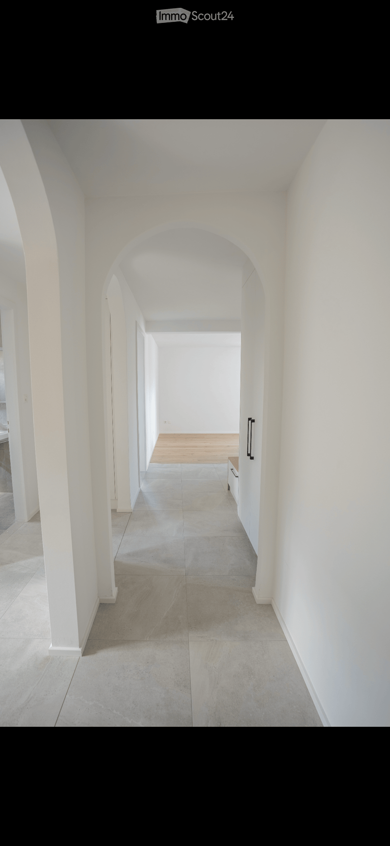 A long, white-walled corridor with arched doorways, polished concrete floors, and minimal furnishings.