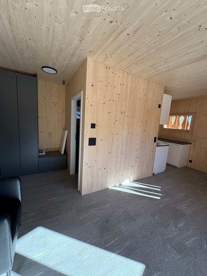 Empty room with wooden walls, wooden flooring, and white cabinets. An entrance door is on the left, with a bench and closets. A small kitchen is on the right with a countertop and window.
