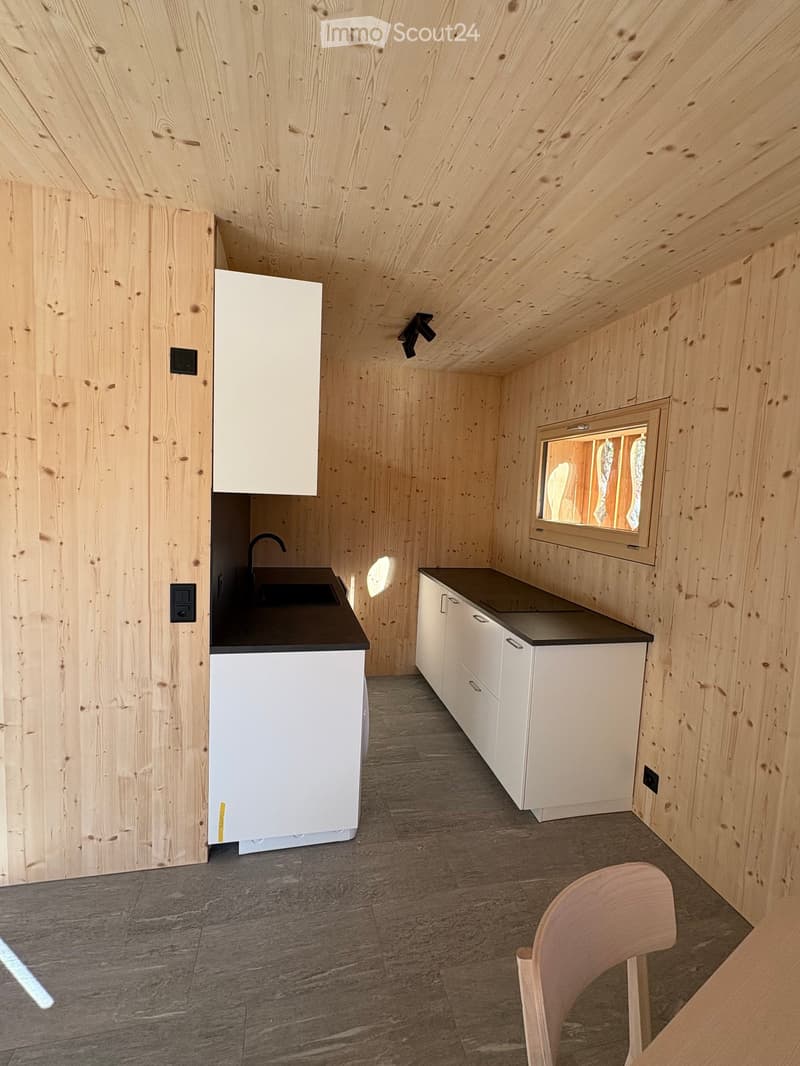 The interior of a kitchen with white cabinets, a black countertop, a sink with a faucet, a window, wooden walls, and a ceiling light.