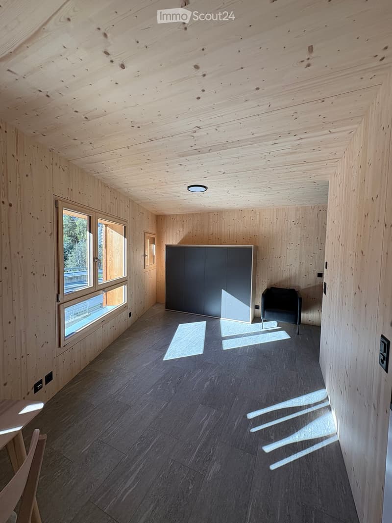 Wooden panel walls and ceiling, large window with a view of the outdoors, grey cupboard, black chair, wooden floor, ceiling lamp