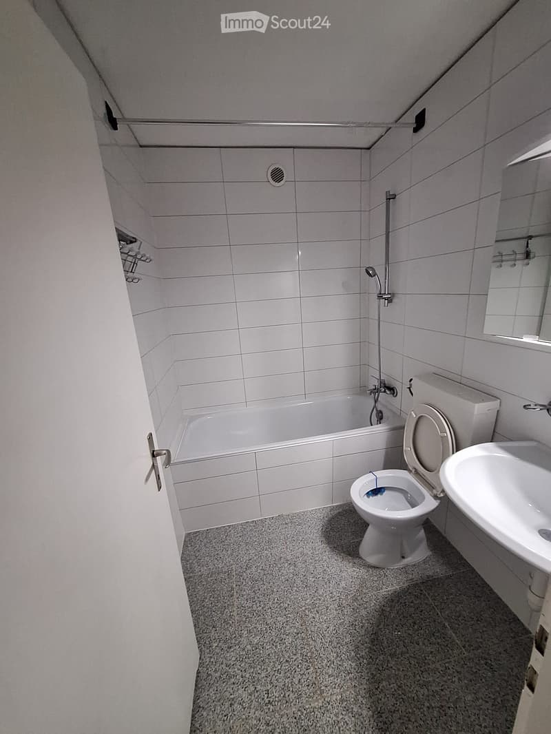 Modern bathroom with white tiled walls, a bathtub, toilet, sink, and mirror. The floor is covered with a speckled gray carpet.