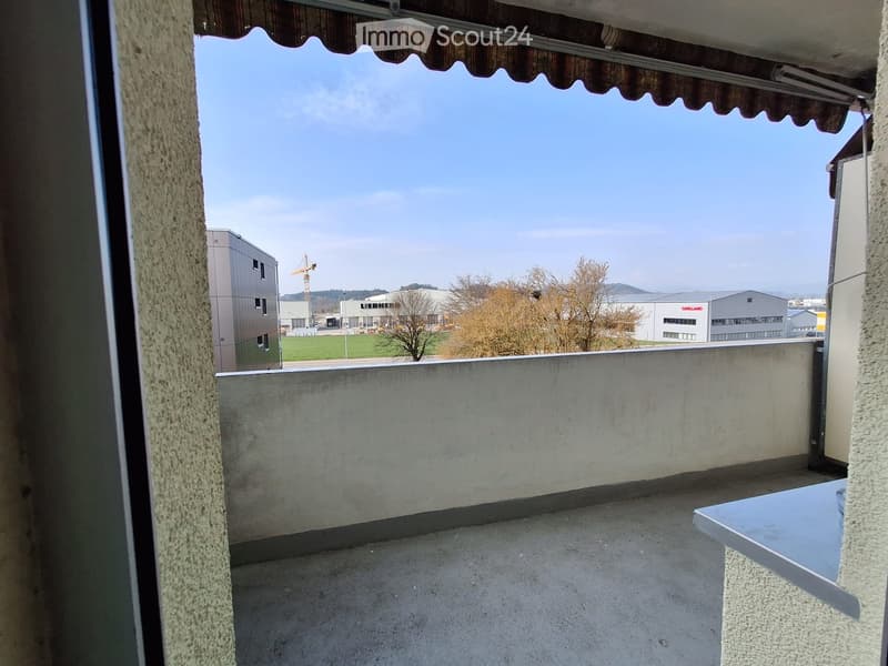 Balcony with concrete floor, low wall, and a small table. View includes industrial buildings and a crane in the distance.