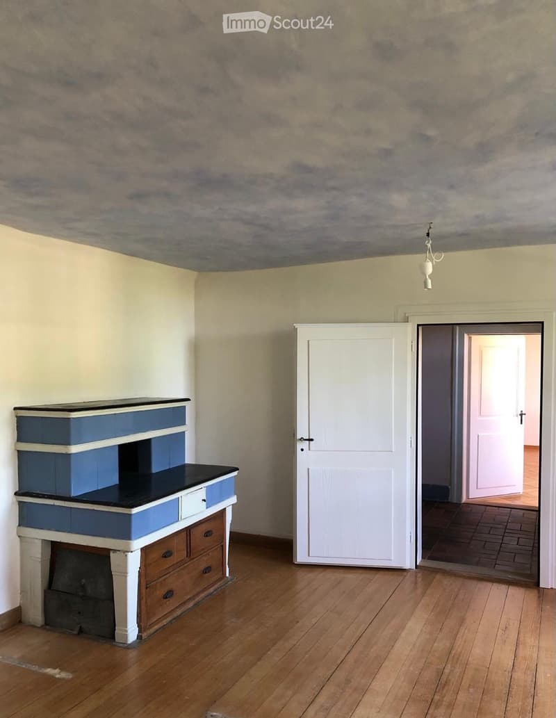An unfurnished room with a fireplace and a door leading to another room. The fireplace has a black top and blue drawers, and the room has wooden flooring and light-colored walls.