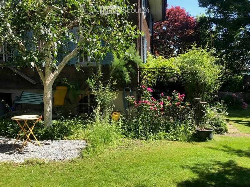 A well-maintained garden with a variety of plants, a small wooden table, a bench, and a tree providing shade. The garden is bordered by a house with visible windows and a balcony.