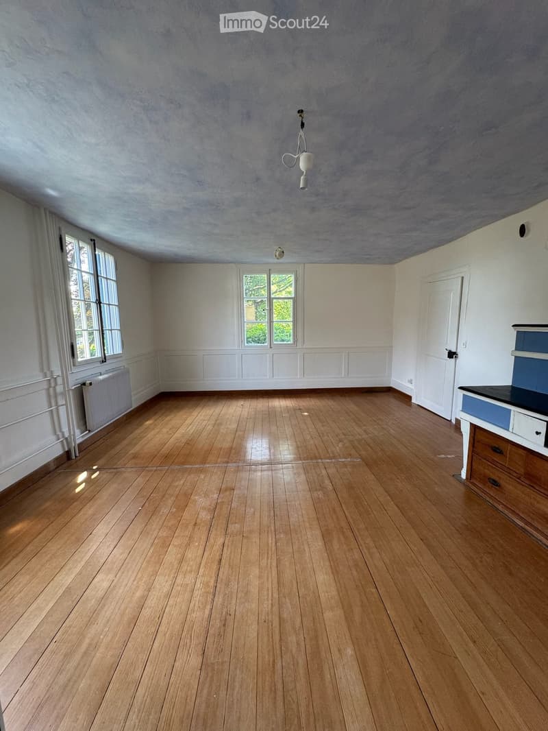 Empty room with wooden flooring, white walls, and large windows. A small cabinet is visible in the corner.