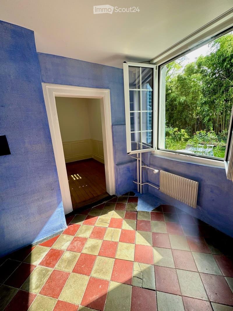 Empty room with blue walls, wooden floor, tiled section, radiator, and large window with view of greenery.