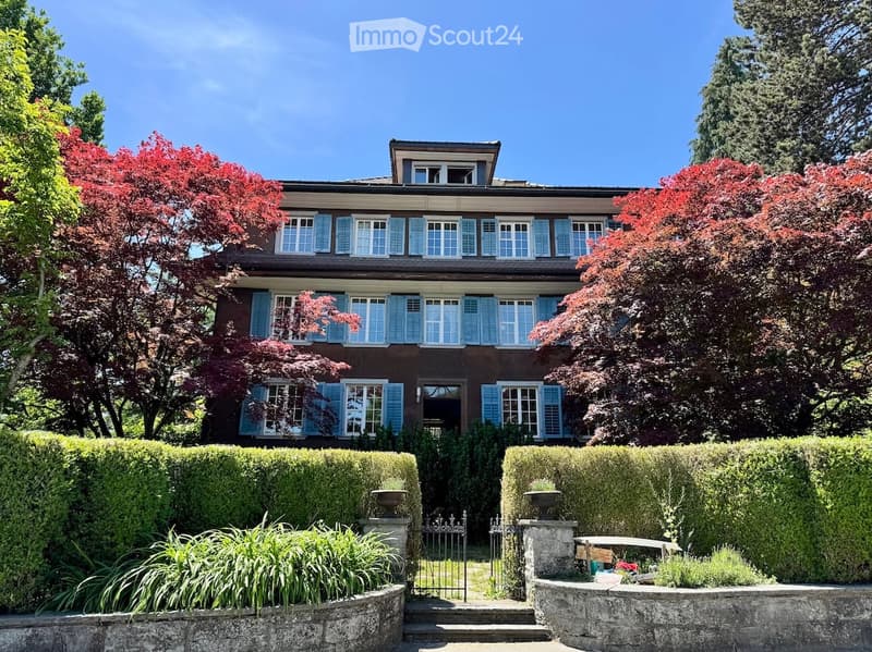 A two-story house with a dark brown exterior and light blue shutters, surrounded by well-maintained hedges and trees.