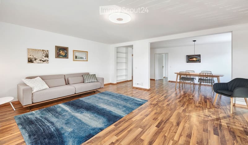 modern living room with wooden floor, gray couch, blue rug, dining table, chairs, white walls, ceiling light, paintings