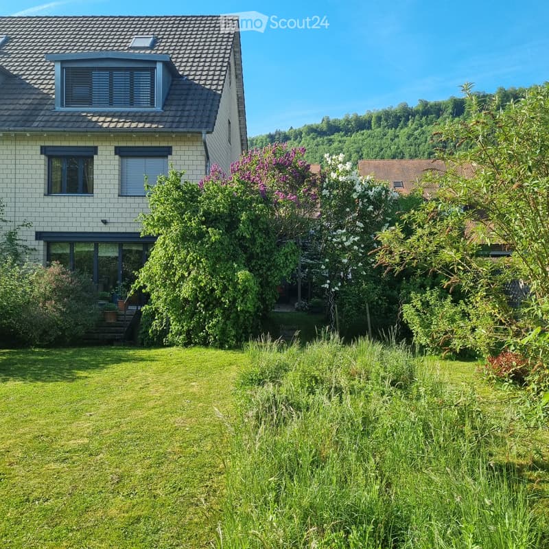 A well-maintained garden with a lawn, various flowering shrubs, and trees, surrounding a two-story house with a tiled roof and multiple windows.