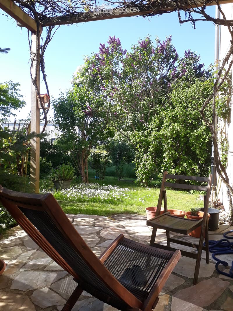 Garden area with stone patio, wooden chairs, potted plants, and lush greenery with flowering trees.