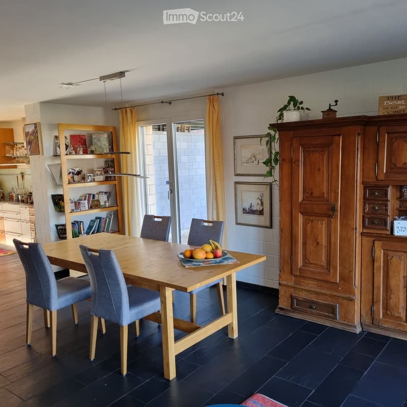 Dining room with wooden table, 4 chairs, wooden cabinet, shelves with books, framed pictures, and a plant on top