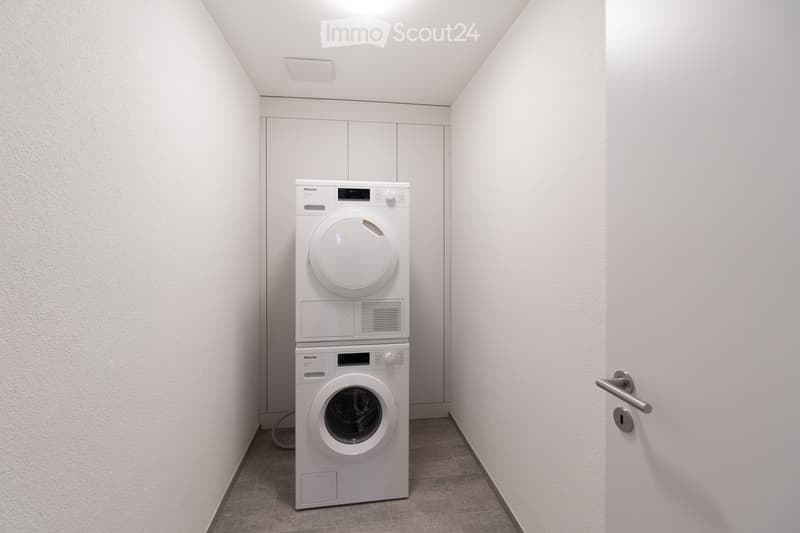 A small room with a white washing machine and dryer stacked on top of each other, white walls, and a white door.