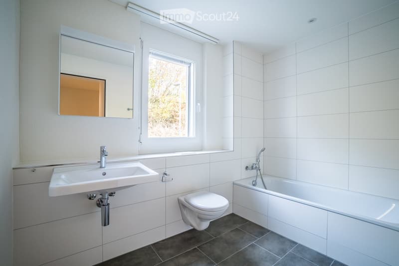 Modern bathroom with white tiles, a sink, a toilet, a bathtub, and a large mirror above the sink. A window provides natural light.