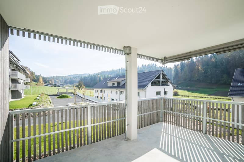 Balcony with wooden railing, overlooking a green landscape with houses and trees.