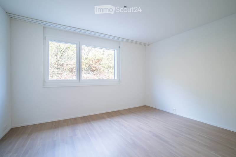 Empty room with white walls, light wooden flooring, and large windows offering a view of the outdoors.