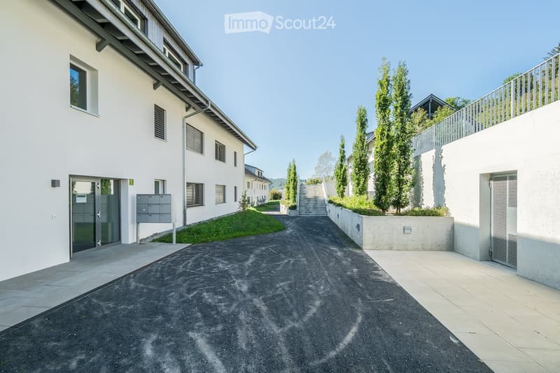 Modern residential building with white facade, multiple windows, and a paved driveway leading to a staircase. The building features a balcony with a metal railing and is surrounded by greenery and trees.