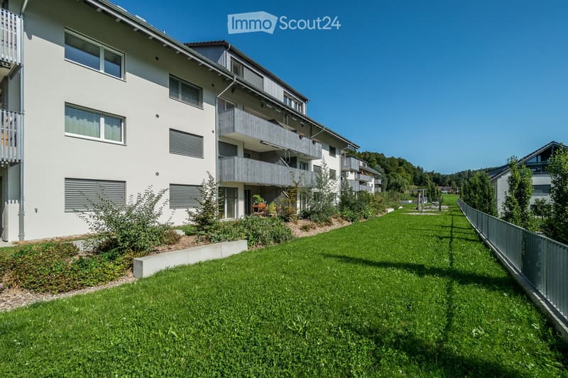 Modern multi-story residential building with white exterior, multiple balconies, and surrounding green lawn.