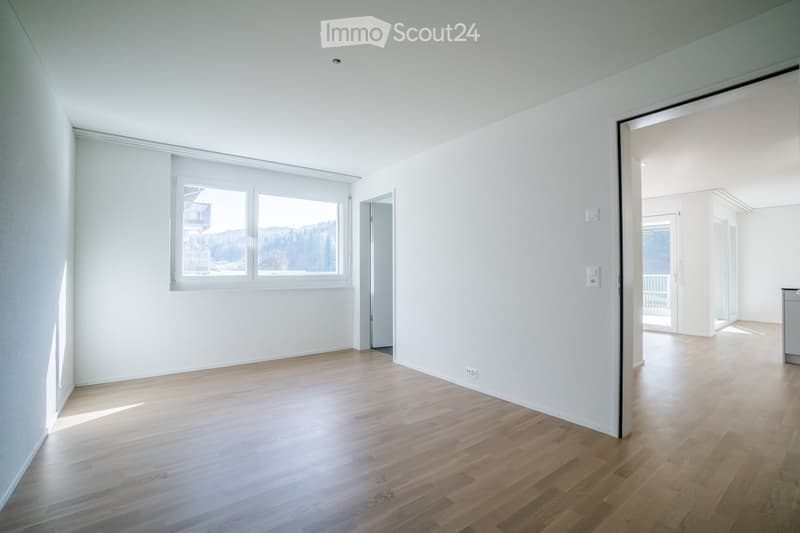 Empty room with light wooden flooring and white walls, featuring large windows and an open doorway leading to another space.