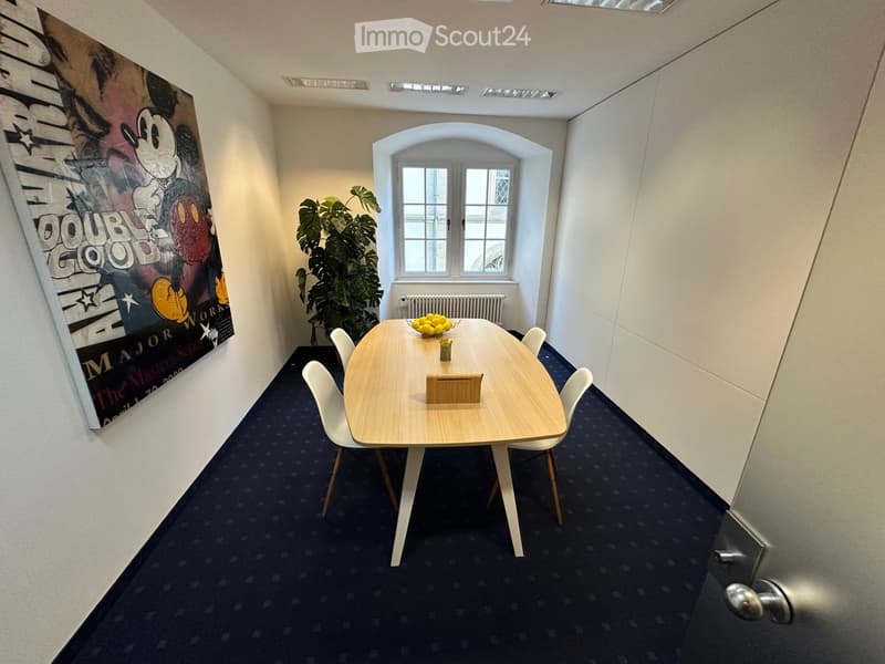 A small, well-lit room with a round wooden table, four white chairs, a bowl of lemons, a small plant, and a large poster on the wall.