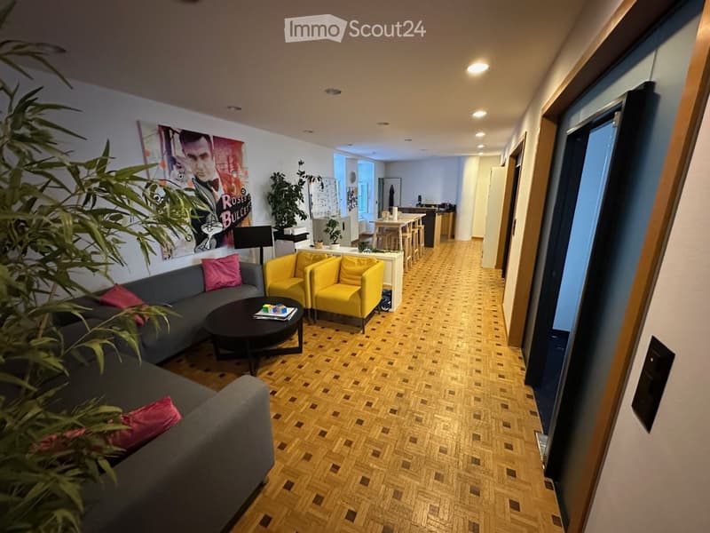Living room with grey sofa, pink cushions, yellow chairs, coffee table, and a poster on the wall. The room has wooden flooring and leads to a kitchen area with a dining table and chairs.