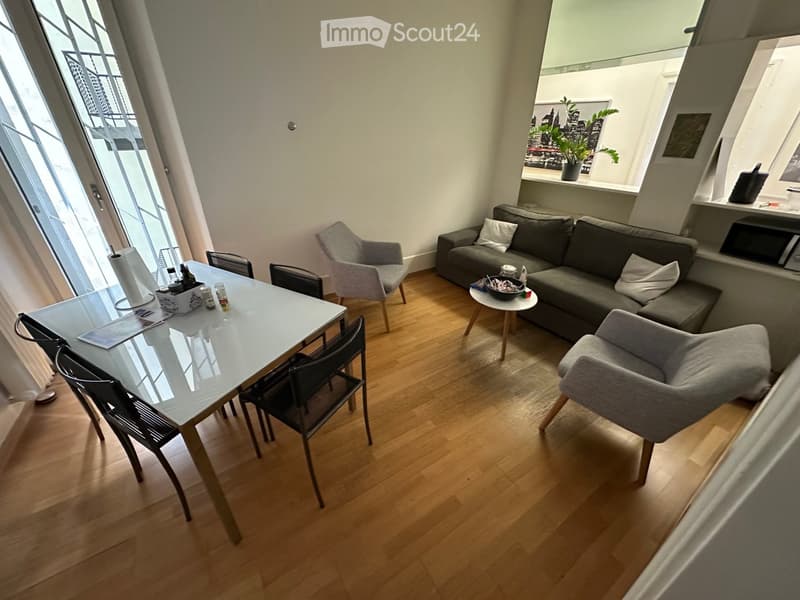 Modern living room with wooden flooring, large windows, grey sofa, chairs, and a dining table.
