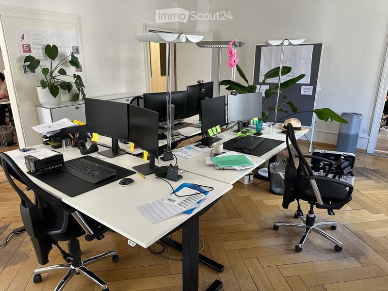 Modern office space with desks, chairs, multiple monitors, laptops, keyboards, and a whiteboard. Wooden flooring and large windows provide natural light.