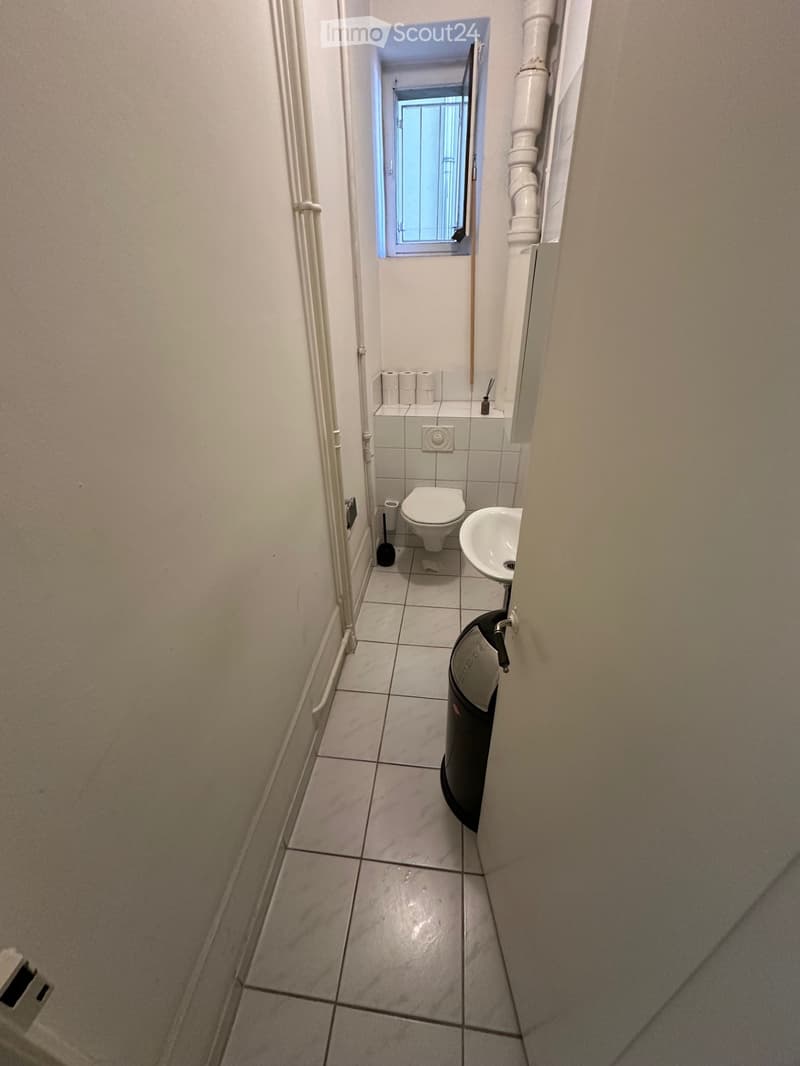 Small bathroom with white tiled floor and walls, toilet, sink, toilet paper, and a trash bin.