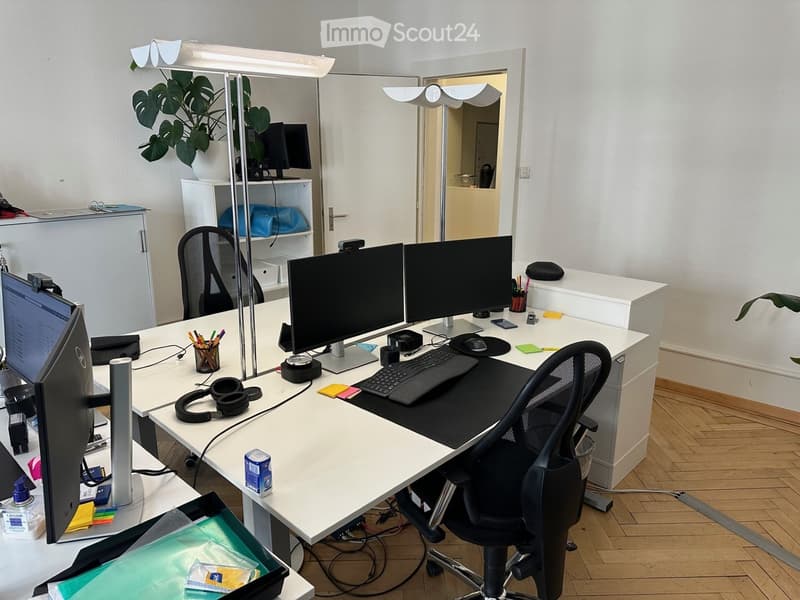 Office space with a large desk, multiple computer monitors, ergonomic chair, and various office supplies including headphones, a plant, and a shelf with additional equipment.