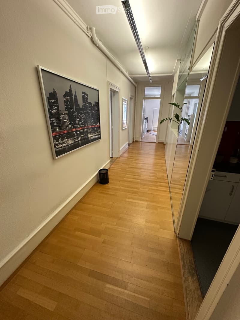 A modern hallway with light wooden flooring, white walls, and a large framed cityscape picture on the left wall. A small black trash can is placed near the picture, and a glass partition on the right leads to another room.