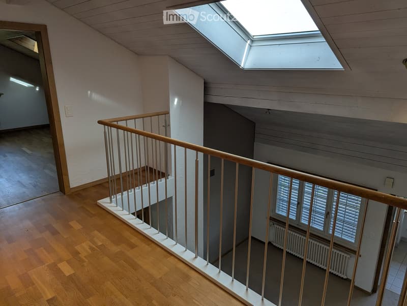Spacious hallway with wooden flooring, white walls, and a modern staircase featuring a wooden handrail and metal balusters. A skylight provides natural light, and a window with blinds is visible in the background.