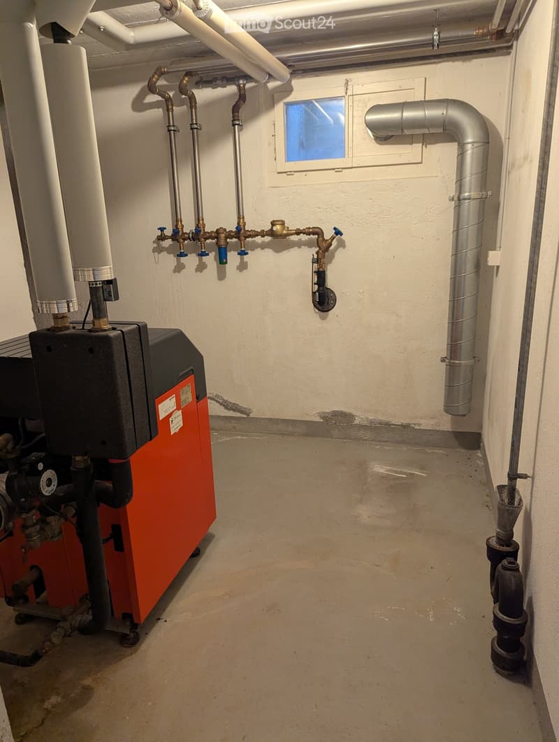 Utility room with heating system, water tank, pipes, and valves. Exposed concrete floor and white walls.
