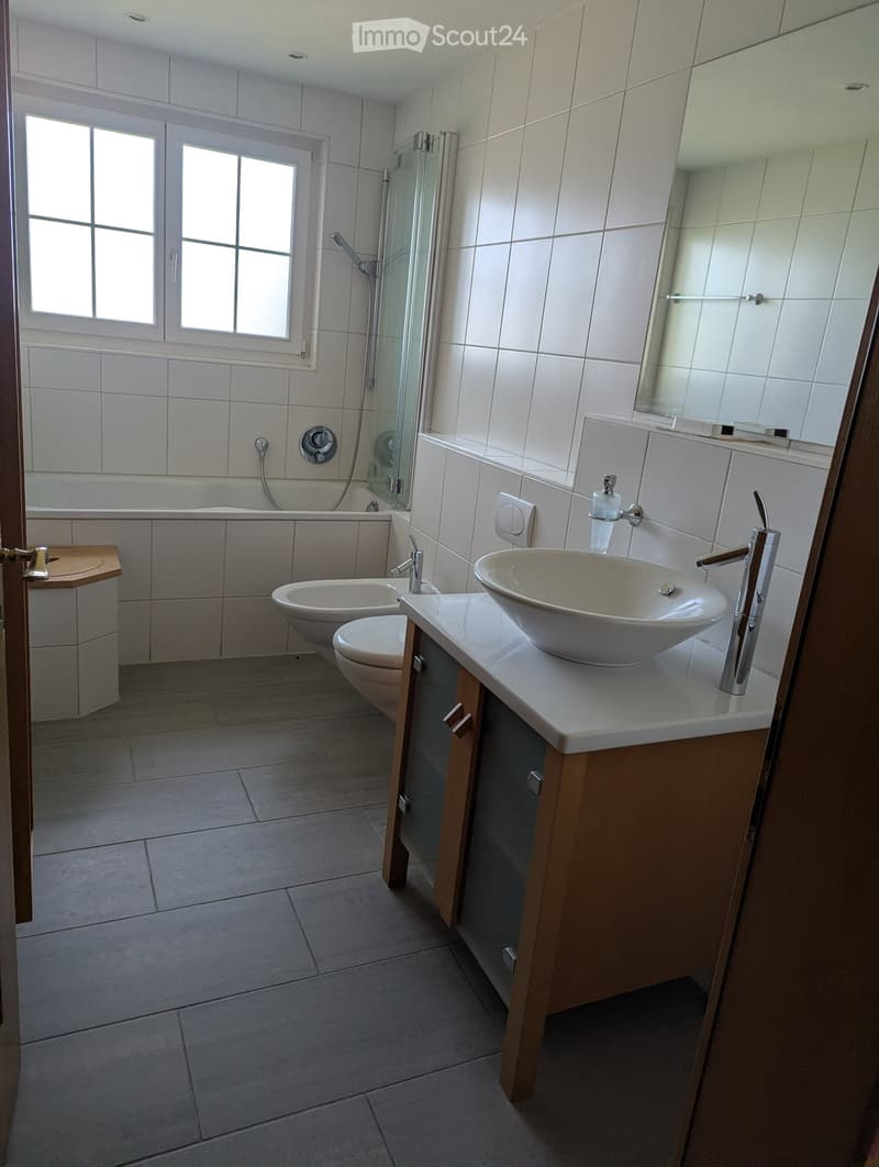 Modern bathroom with white tiles, a large sink with a modern faucet, a toilet, a bathtub, and a glass-enclosed shower area. The room features a large mirror above the sink and a window providing natural light.
