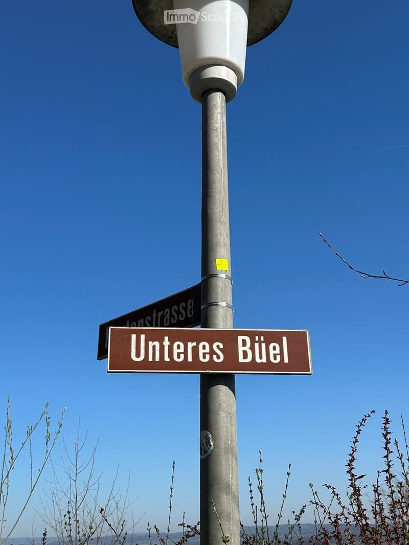 Street sign reading "Unteres Büel" mounted on a pole against a clear blue sky, with sparse vegetation in the background.