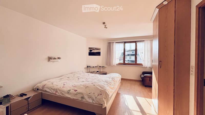 Bedroom with bed, wooden floor, white walls, window with curtains, wooden cabinet, small table, picture frame, and a brown armchair.