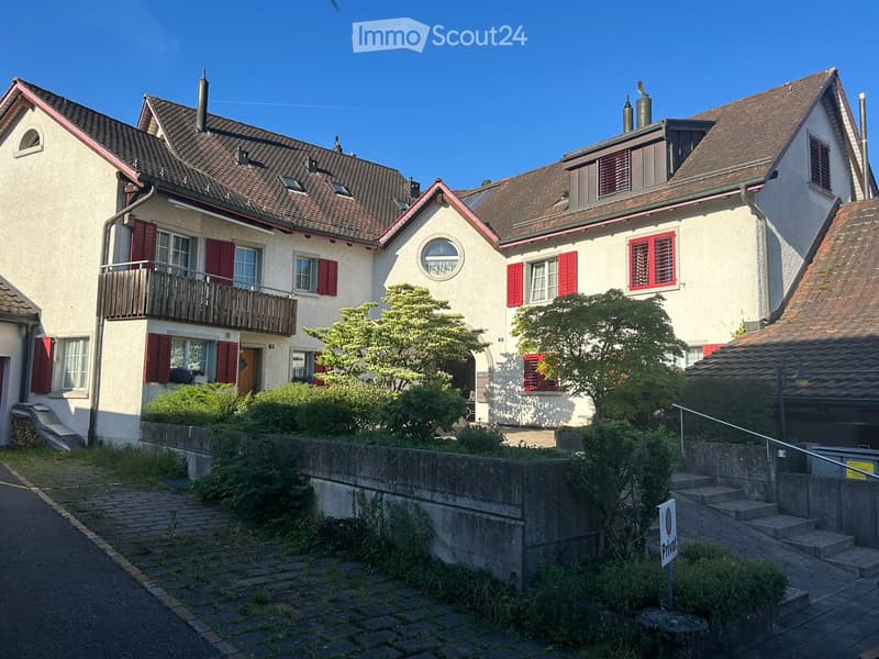 Two-story house with red shutters, white walls, and a balcony on the upper floor. The house has a tiled roof and is surrounded by greenery and a low stone wall.