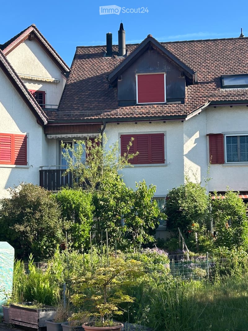 A two-story house with a tiled roof, white exterior walls, and red shutters on the windows. The house features a small balcony on the left side and is surrounded by lush greenery and potted plants in the front yard.