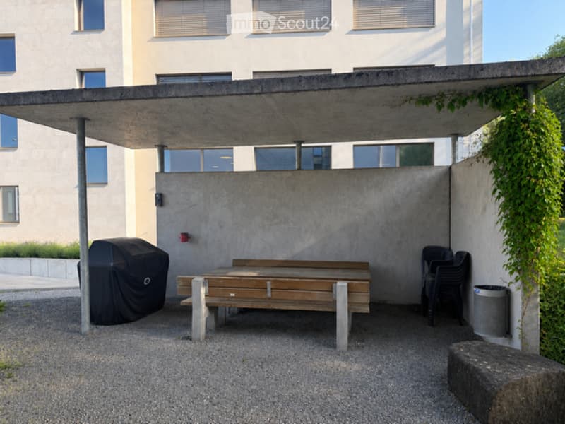 Modern outdoor seating area with a wooden bench, covered by a concrete roof, surrounded by a concrete wall and greenery. Includes a covered grill and plastic chairs.