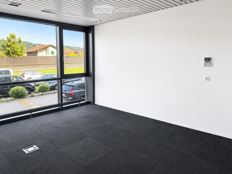 Empty room with white walls, grey carpet, large glass sliding doors, and a view of a parking lot and greenery.