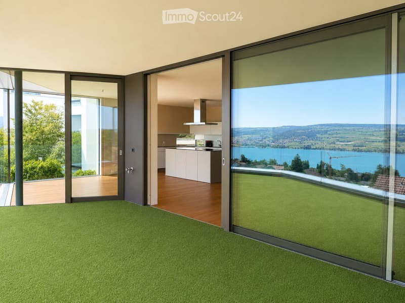 Bright, modern room with green carpeting, large glass sliding doors opening to a balcony, and a visible kitchen area with white cabinets and stainless steel appliances.