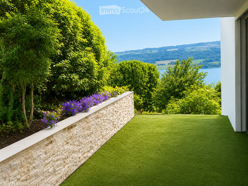 Balcony with artificial grass, stone low wall, purple flowers, and scenic countryside view.