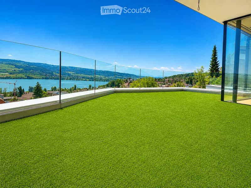 A modern rooftop terrace with artificial grass, glass railing, and scenic lake and mountain view.