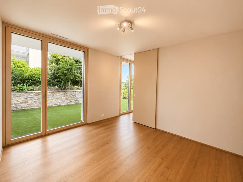 Empty room with wooden flooring, large sliding glass doors leading to a garden, and neutral-colored walls.