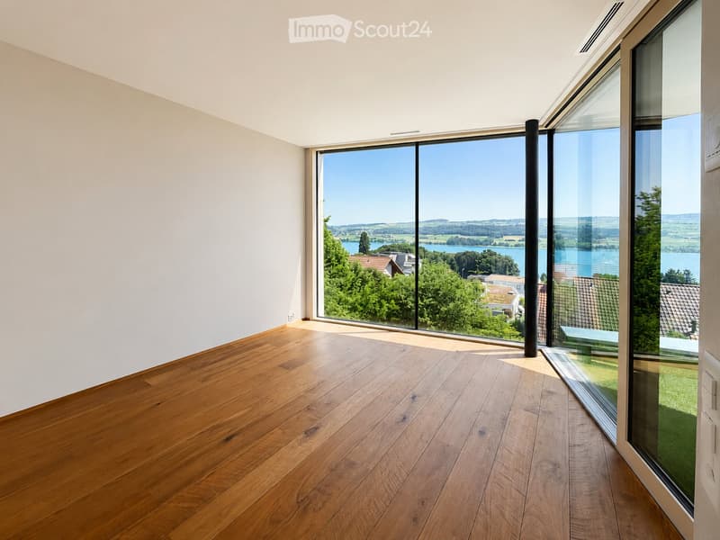 Empty room with wooden flooring and large glass windows offering a scenic view of a lake and surrounding landscape.