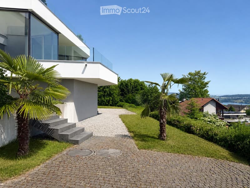 Modern two-story house with white exterior, large glass windows, and a raised ground floor. The entrance features concrete steps leading up to the house, surrounded by palm trees and a well-maintained garden with a gravel pathway.