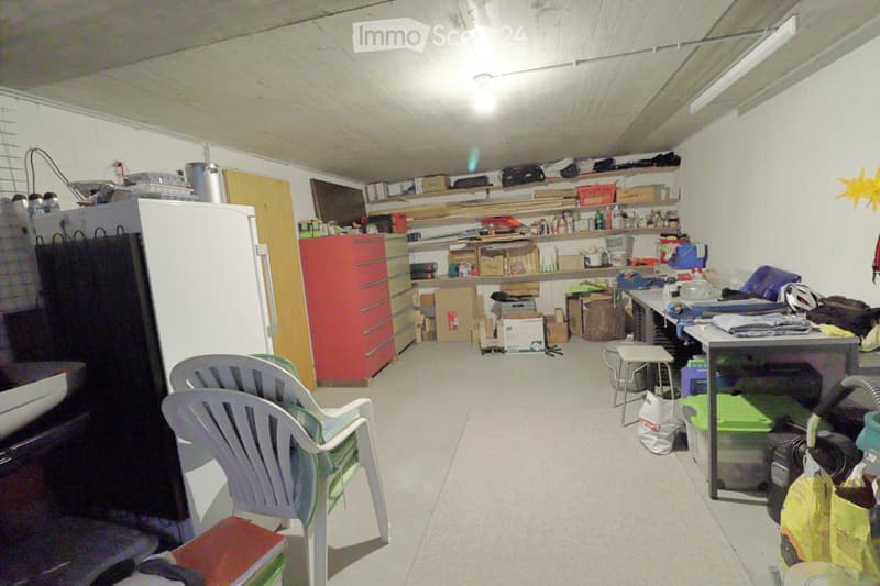 A cluttered storage or utility room with plastic chairs, a small table, shelves filled with various items, a refrigerator, and a red drawer unit.
