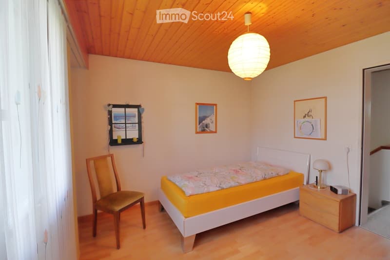 Cozy bedroom with wooden floor, slanted wooden ceiling, bed with yellow bedspread, wooden chair, nightstand with lamp, and framed pictures on the wall.