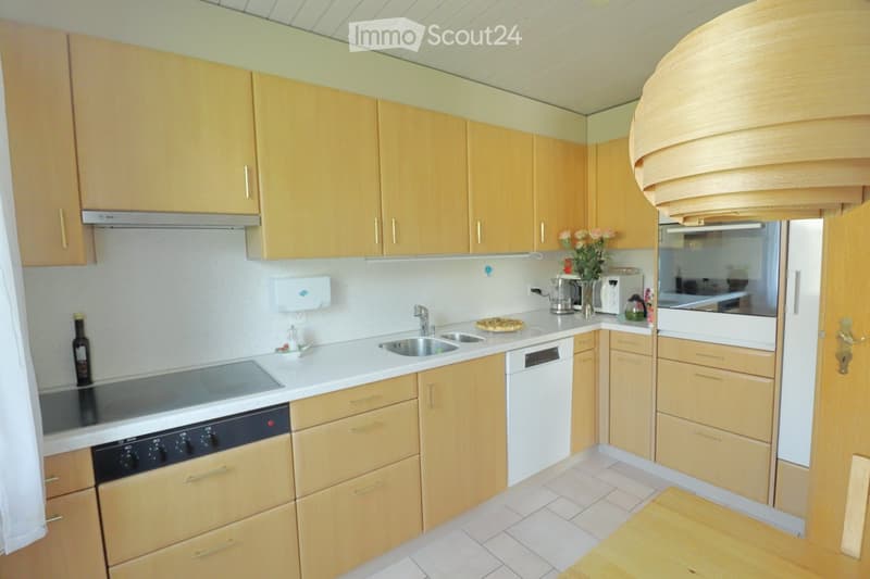 Bright kitchen with light wooden cabinets, white countertops, stainless steel sink, oven, dishwasher, and various appliances. A pizza sits on the counter, and a vase with flowers is visible.